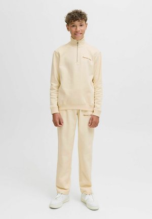 Vaaleanbeige vetoketjullinen huppari, jossa on korkea kaulus ja yhdistelevät verkkarit. Pehmeä rakenne, ribbaus hihansuissa ja brodeerattu logo. Valkeat lenkkarit.