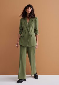 Conjunto de traje verde oliva que incluye un blazer de doble botonadura y pantalones de pierna ancha, complementado con zapatos negros de suela gruesa sobre un fondo texturizado.