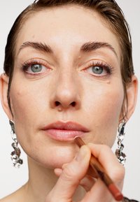 Una persona con la pelle chiara e orecchini d'argento applica una matita per labbra nude sulle proprie labbra su uno sfondo bianco semplice.