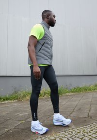 Sportig outfit med en grå puffert väst, en ljusgrön kortärmad skjorta, svarta leggings och ljusblå sportskor med detaljer.