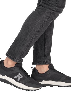 Persoon draagt zwarte en grijze sneakers met witte zolen en zwarte spijkerbroek, benen gekruist bij de enkels, op een witte achtergrond.