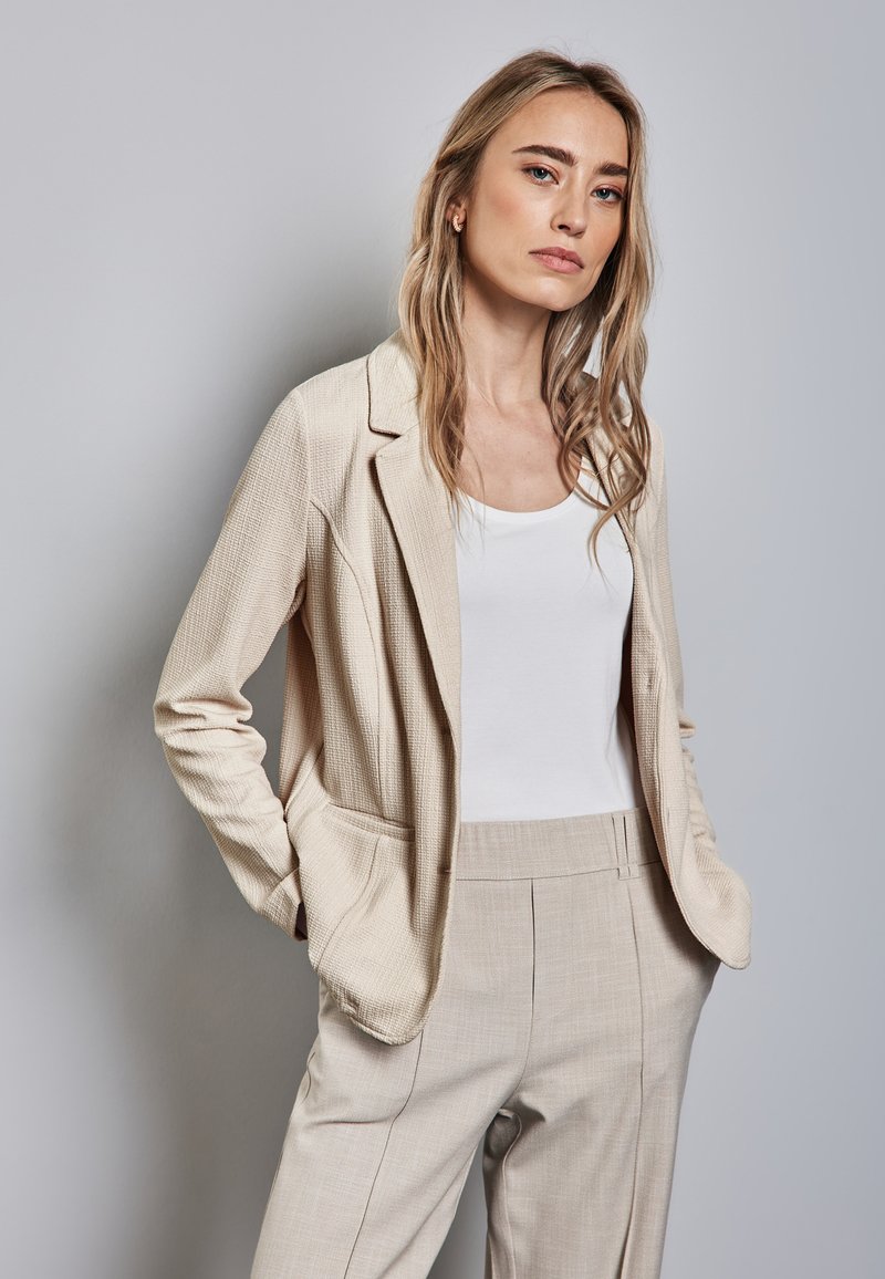 Harmaa teksturoitu bleiseri löysällä mitalla, jossa on taskut ja lovikaulus, yhdistetty valkoiseen hihattomaan paitaan ja harmaisiin räätälöityihin housuihin.