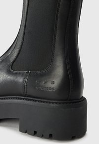 Bottines noires avec une tige en cuir lisse et un panneau élastique, dotées d'une semelle en caoutchouc robuste et d'un logo en relief.