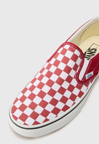 Zapatilla de lona tipo slip-on a cuadros rojos y blancos con suela de goma blanca y costuras rojas en contraste. Presenta una etiqueta con el logo de Vans en el lateral.