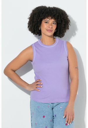 Femme aux cheveux bouclés portant un haut lavande sans manches et un jean bleu clair avec des patchs étoiles multicolores, debout la main sur la hanche.