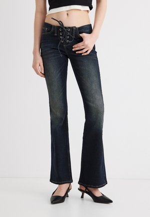 Spodní část těla osoby nosící černé džíny s rozšířenými nohavicemi na šněrování a černé lodičky s podpatkem, stojící na jednobarevném bílém pozadí.