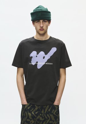 Junger Mann trägt ein dunkles T-Shirt mit lila abstraktem Logo und Text, grüne Mütze und camouflagegemusterte Hose, steht mit den Händen in den Taschen.