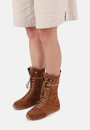 Jambes portant une jupe beige et des bottes montantes en daim marron à lacets avec semelles plates, debout sur un fond blanc.