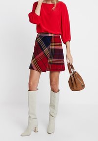 Rode blouse met pofmouwen, geruite minirok in rood, blauw en geel, grijze kniehoge laarzen en een bruine handtas.