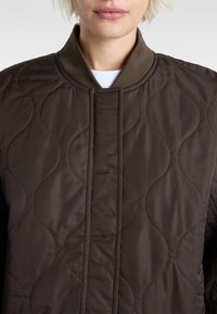 Braune gesteppte Bomberjacke mit ribbeltem Kragen. Verfügt über ein wellenförmiges Stickmuster und einen Frontverschluss ohne sichtbare Knöpfe oder Verschlüsse.
