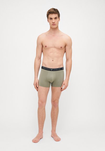 Boxeri scurți de culoare verde măslinie, fabricați dintr-un material elastic, cu un brâu negru pe care este inscriptionat logo-ul brandului, design ajustat și lungime până la mijlocul coapselor.