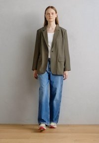 Blazer khaki con un bottone singolo, due tasche frontali, indossato sopra una maglietta bianca e jeans leggermente blu e larghi. Sneakers casual completano l'outfit.