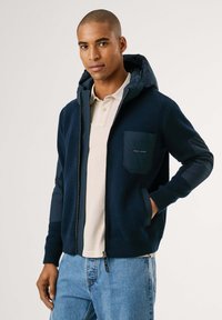 Hombre que lleva una sudadera con cremallera azul marino y capucha, con bolsillo en el pecho, sobre una camiseta de polo beige y jeans azules, de pie frente a un fondo liso.