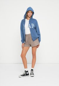Blå hettegenser med hvit "GAP" tekst, kombinert med lysegrå shorts. Modellen har på seg svarte høyhælte sneakers og hvite sokker.