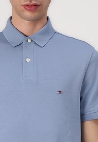 Polo di colore blu chiaro realizzato in tessuto strutturato, con colletto classico e due bottoni, dotato di un piccolo logo in rosso, bianco e blu.