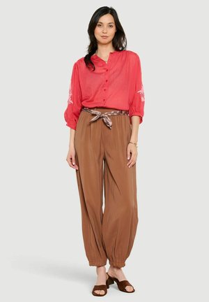 Žena stojaca oblečená v červenej blúzke so zapínaním na gombíky, hnedých vysokých nohaviciach s pásikom a hnedých topánkach na blokovom podpätku s otvorenou špičkou.