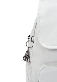 Mochila de tela blanca con cierre de solapa, superficie texturizada y bolsillos con cremallera, con un charm de robot metálico adjunto al tirador de la cremallera.