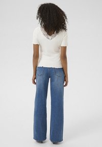 Weißes, geripptes T-Shirt mit Spitzen-V-Ausschnitt, kombiniert mit hoch taillierten, weiten blauen Jeans; mit gewelltem Saum am Oberteil.