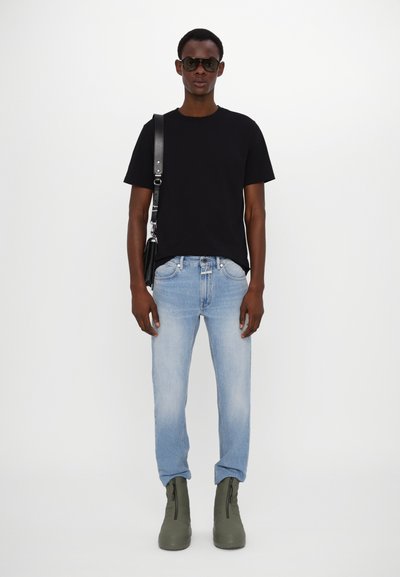 T-shirt noir à col rond, jeans droits bleu clair, bottines grises à la cheville et sac bandoulière noir ; modèle portant des lunettes de soleil foncées.