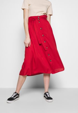 Femme portant un haut beige, une jupe midi rouge avec des boutons et une ceinture, ainsi que des baskets noires, debout devant un fond clair uni.