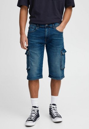 Hombre con pantalones cortos cargo de mezclilla azul, zapatillas negras de caña alta con cordones blancos, calcetas blancas y una camisa oscura, de pie con una mano en el bolsillo.