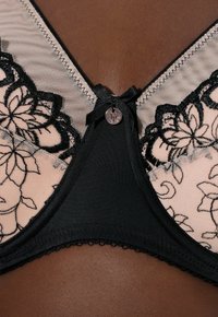 Sujetador de encaje negro con bordado floral transparente, un lazo de satén y detalles metálicos en el centro, con una banda de tela negra suave y bordes con ondas.