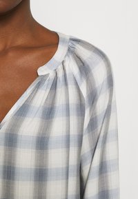Blouse à carreaux bleu clair et blanc avec un col décontracté, des détails froncés aux épaules, et un tissu doux et texturé.