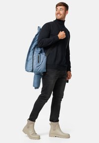 Lyseblå dunryggsekk med svarte stropper, båret av en person i svart genser med høy hals og svarte jeans, komplementert av beige gummistøvler.