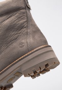 Harmaa nahkasaapas, jossa on teksturoitu pinta, erottuva saumaus, kuminen pohja ja puulogo-koriste. Käytännöllinen yläosan muotoilu tarjoaa tukea.