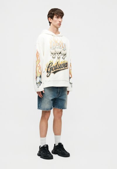 Noor mees valge kapuutsiga, millel on leegitsevad koljud ja "Gcds" tekst, teksapüksid, valged sokid ja mustad tossud, seisab ühevärvilise taustaga.