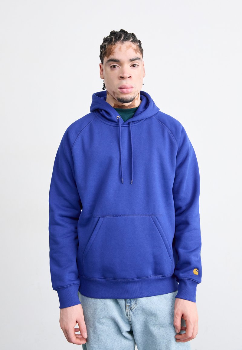 Blue hoodie with a kangaroo pocket, drawstring hood, ribbed cuffs, and hem. Made from smooth fabric, featuring a logo on the left cuff.