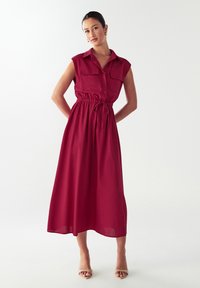 Burgundin värinen hihaton mekko, jossa on nappisuljenta edessä, kaksi rintataskua, vyötäröltä kireä malli ja virtaava puolipitkä hameosa. Silkkinen kangas.