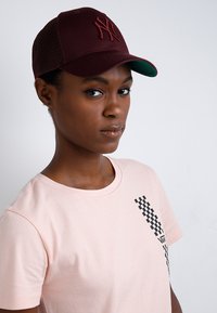 Cappellino da baseball color bordeaux con visiera verde e logo ricamato, abbinato a una t-shirt leggera rosa chiaro con un design grafico a scacchi neri.