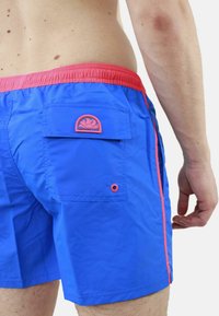 Pantaloni da nuoto blu con una vita rossa e accenti laterali. Presentano una tasca sul retro con un logo a onda e un piccolo dettaglio con occhiello.