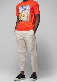 Pomarańczowy bawełniany T-shirt z fotograficznym nadrukiem, zestawiony z jasnobeżowymi chinosami i czarnymi trampkami z białymi podeszwami.