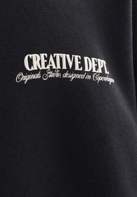 Black fabric apparel featuring white embroidered text that reads "CREATIVE DEPT. Originals Studio, designed in Copenhagen."