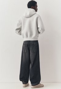 Personne portant un sweat à capuche gris clair et un jean noir large, debout face à un mur blanc, les mains sur les hanches, chaussée de baskets beiges.