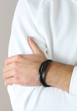 Schwarzes Lederarmband mit zwei geflochtenen Strängen und einem glatten Akzentstück, das an einem Handgelenk zu einem strukturierten weißen Hemd getragen wird.