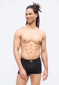 Γελαστός άνδρας χωρίς πουκάμισο με ντρέντλοκς, φορώντας μαύρα μπόξερ Calvin Klein, στέκεται μπροστά σε απλό ανοιχτό φόντο.