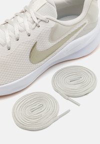 Světlé béžové sportovní boty s síťovinovým svrškem, ikonickým logem Nike v matném zlatě a plochými tkaničkami. Obsahuje dvě světle šedé kulaté tkaničky na spodní části.