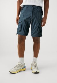 Mörk teal cargo shorts med flera sidofickor, knappstängning och avslappnad passform, tillverkade av lättviktig tyg. Bärs med sneakers.