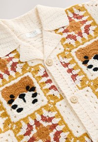 Virkat kofta med en strukturerad krämfärgad bas och bruna, orange och svarta partier. Den har en krage och träknappar.