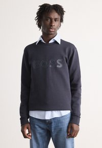 Σκούφος ναυτικού χρώματος με υφή λογότυπο "BOSS", στρογγυλή λαιμόκοψη και ριμπ μανσέτες, φορεμένος πάνω από ένα φωτεινό μπλε πουκάμισο με γιακά και τζιν παντελόνι.