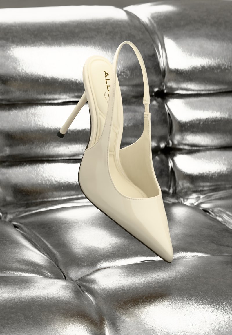 Chaussure à talon haut slingback en cuir verni crème, pointe pointue et talon fin, posée sur une surface texturée argentée brillante.