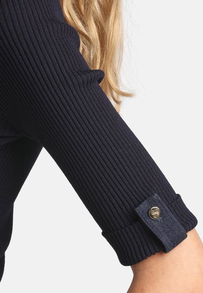 Gerippter Ärmel aus marineblauem Stoff mit Jeansmanschette, verziert mit einem dekorativen goldfarbenen Knopf. Glatte Textur mit enger Passform.