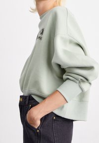 Ljust grön bomullströja med en kortad design, vida ärmar och ribbade mudd; har en kontrasterande grafik på framsidan.