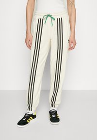 Pantaloni della tuta color crema con strisce laterali nere, vita elastica e cordoncino verde, abbinati a sneakers nere con strisce gialle.