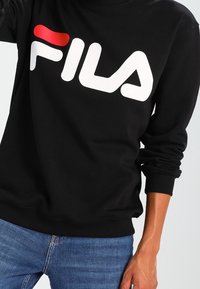 Sudadera negra con un destacado logo blanco de "FILA" y un acento rojo. Fabricada en un tejido suave, con puños y dobladillo acanalados, de corte casual.