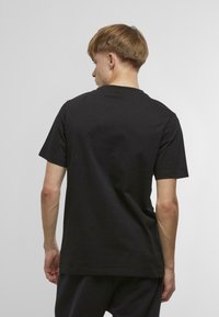 Melna kokvilnas T-krekls ar klasisku apaļu apkakli un īsām piedurknēm, skatīts no aizmugures. Gluda tekstūra, vienkāršs dizains un bez redzamām zīmēm.