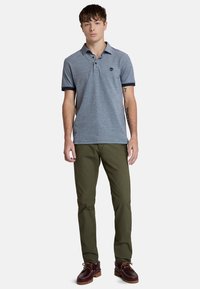 Tummansininen poolopaita lyhyillä hihoilla, jossa on kaulus ja logo. Yhdistetty oliivinvihreisiin housuihin ja ruskeisiin venepolkimiin.